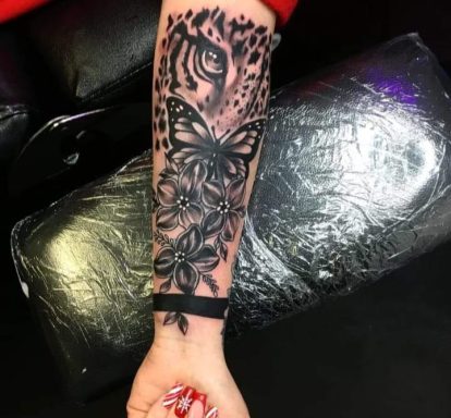 Black and grey tattoo featuring a butterfly, floral designs, and intricate detailing on a forearm.