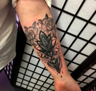 Black and grey tattoo featuring a lotus flower and intricate floral patterns on the forearm.