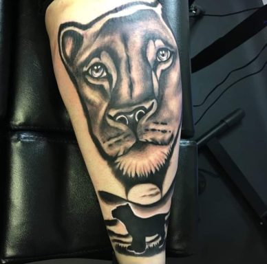 A detailed black and grey tattoo of a lioness's face on a person's leg.