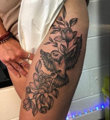 Tattoo of a bird flying among flowers on a person's thigh.