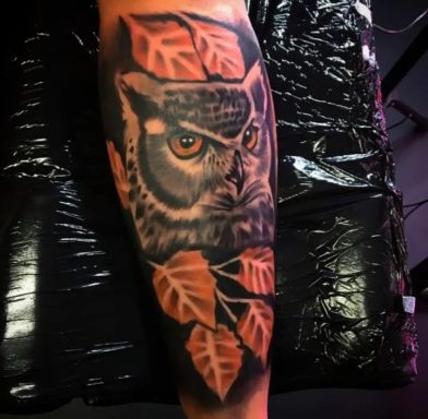Tattoo of an owl surrounded by autumn leaves on a leg.