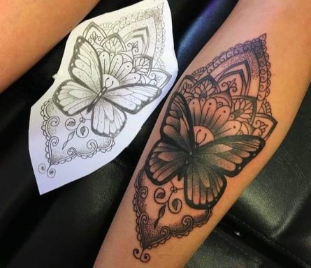 Tattoo of a butterfly surrounded by intricate lace patterns on a forearm, alongside a stencil.