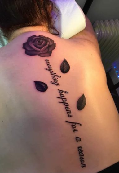 A tattoo on a back featuring a rose and the phrase “everything happens for a reason.”